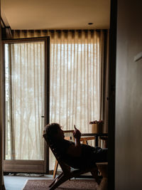 Side view of silhouette of anonymous person sitting in armchair and browsing mobile phone while chilling in room of house at weekend person