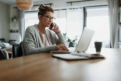 Woman talking over smart phone using laptop on table while sitting at home