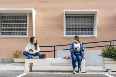 Female friends social distancing wearing protective face mask sitting on concrete bench