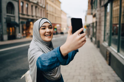 Cheerful young muslim woman wearing hijab taking selfie on sidewalk in city