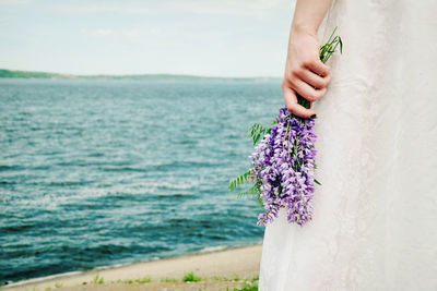 Midsection of bride holding lavenders on shore against sky