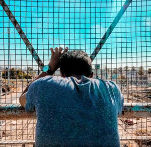 Rear view of man standing by chainlink fence against sky