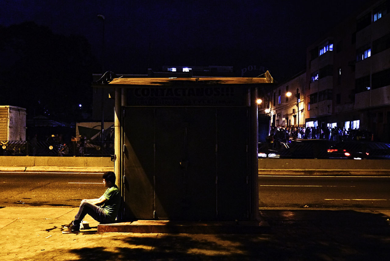 night, architecture, city, street, building exterior, illuminated, built structure, sitting, one person, transportation, real people, road, social issues, nature, city life, men, sidewalk, outdoors