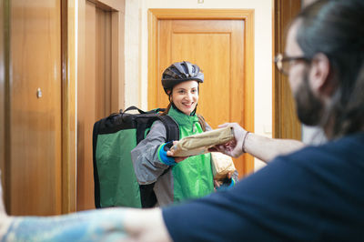 Client receiving paper bag with food from glad delivery woman with thermal backpack through apartment door
