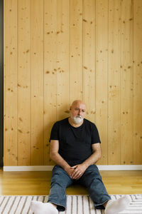 Upset mature man sitting down near wooden wall at home
