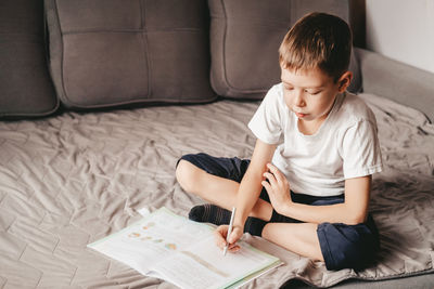 Boy does homework while sitting on the gray sofa. a caucasian teenager writes in a workbook 
