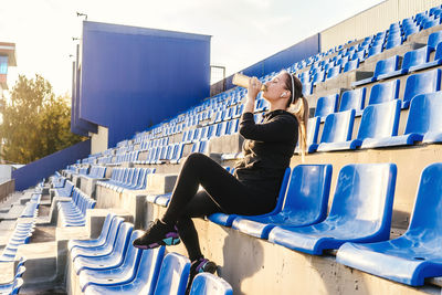 Woman drinking water while sitting on chair in stadium