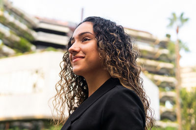 From below side view self assured young hispanic businesswoman in stylish black jacket with curly hair looking away on blurred background of city street