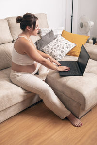 A young pregnant woman works with a laptop on her sofa at home.