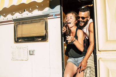 Cheerful couple holding beer bottles standing at entrance of motor home