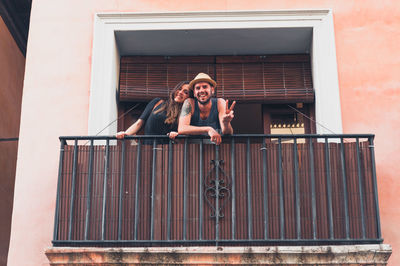 Portrait of smiling young man showing peace sign by girlfriend at balcony