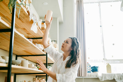 Young woman manager of pottery workshop arranges products on shelf