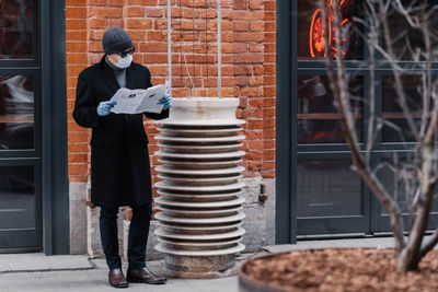 Full length of man wearing mask reading newspaper while standing against wall