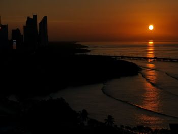 Scenic view of sea by silhouette buildings against sky during sunset