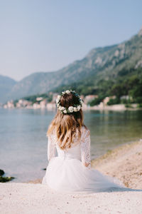 Rear view of bride sitting on sand by bay against sky