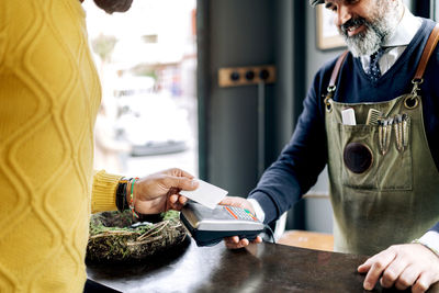 Crop male customer making payment for hairstyle with plastic card on nfc terminal in hands of barber in grooming salon