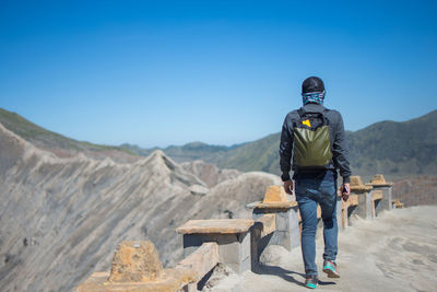 Rear view of backpack man walking on mountain road against clear blue sky