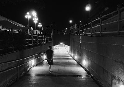 Rear view of woman walking on illuminated footpath by wall at night