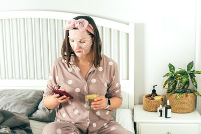 Woman with face mask using mobile phone while sitting on bed at home