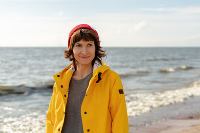 Waist portrait of mature woman in bright yellow cloak and red hat on shore of north sea