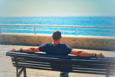 Rear view of man sitting on bench against sea and sky