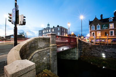 Illuminated street by canal against sky in city