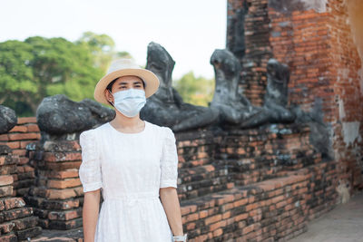 Mid adult woman wearing mask looking away while standing against ancient structure