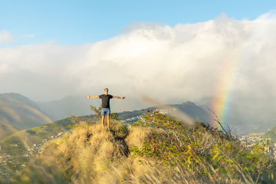 Male hiker with arms outstretched standing on mountain peak against rainbow and cloudy sky