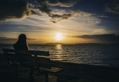 Silhouette woman sitting on bench at beach against sky during sunset