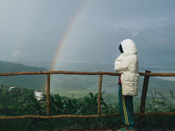 Rear view of man standing on railing against rainbow