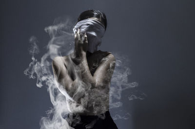 Shirtless woman covering face standing amidst smoke against gray background