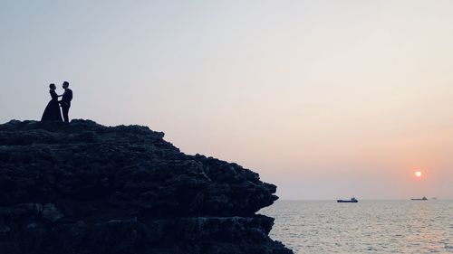 People standing on rock by sea against sky during sunset