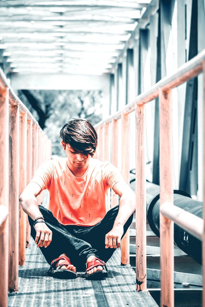 real people, one person, full length, sitting, railing, casual clothing, lifestyles, front view, architecture, day, indoors, built structure, leisure activity, young adult, focus on foreground, staircase, wood - material, looking