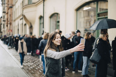 Smiling girl taking selfie through smart phone against crowd in city