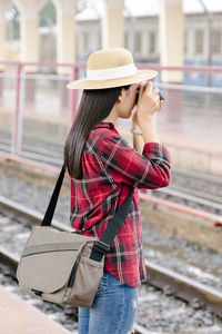 Side view of woman wearing hat holding camera while standing outdoors