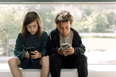 Male and female students using mobile phone while sitting against glass window in school