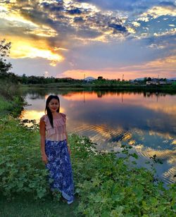 Portrait of smiling woman standing by lake against cloudy sky during sunset
