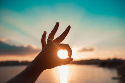 Cropped hand of woman showing ok sign against sky during sunset