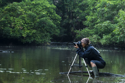 Side view of man photographing lake against trees