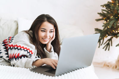 Young asian woman with long hair in cozy sweater using laptop on bed in room with christmas tree 