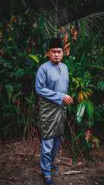 Portrait of young man standing against plants wearing a traditional malaysian malay dress