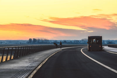 People on sidewalk with vehicle on bridge against sky during sunset