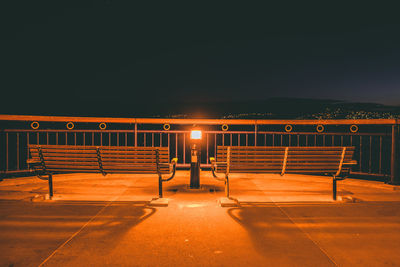 Empty bench by illuminated railing against sky at night