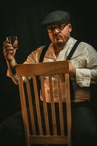 Man wearing hat sitting on chair against black background holding a glass in the hand 