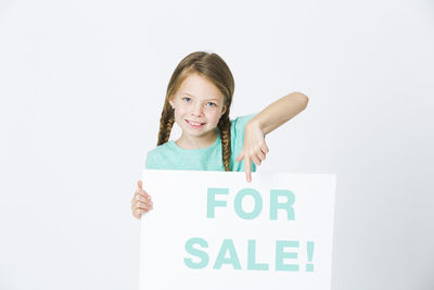 Portrait of cute girl pointing at for sale placard against white background