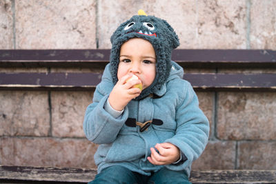 Portrait of cute baby boy eating fruit while sitting on bench against wall