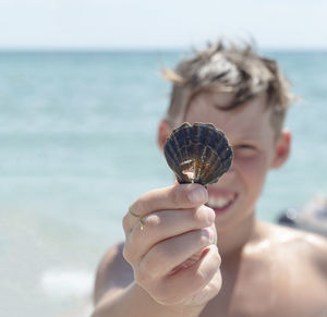 The boy holds a seashell in his hand. the face of a teenager against  the sea. smile.