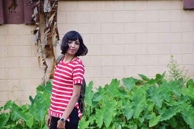 Young woman looking away while standing against plants and wall