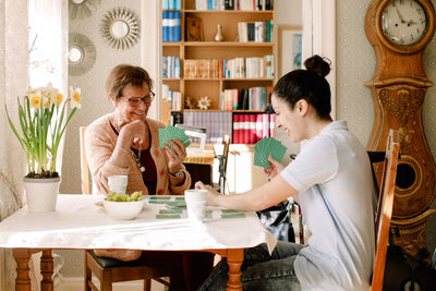 Smiling young female volunteer playing cards with retired elderly woman at table in nursing home