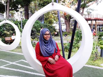 Portrait of smiling young woman wearing hijab sitting on swing in park